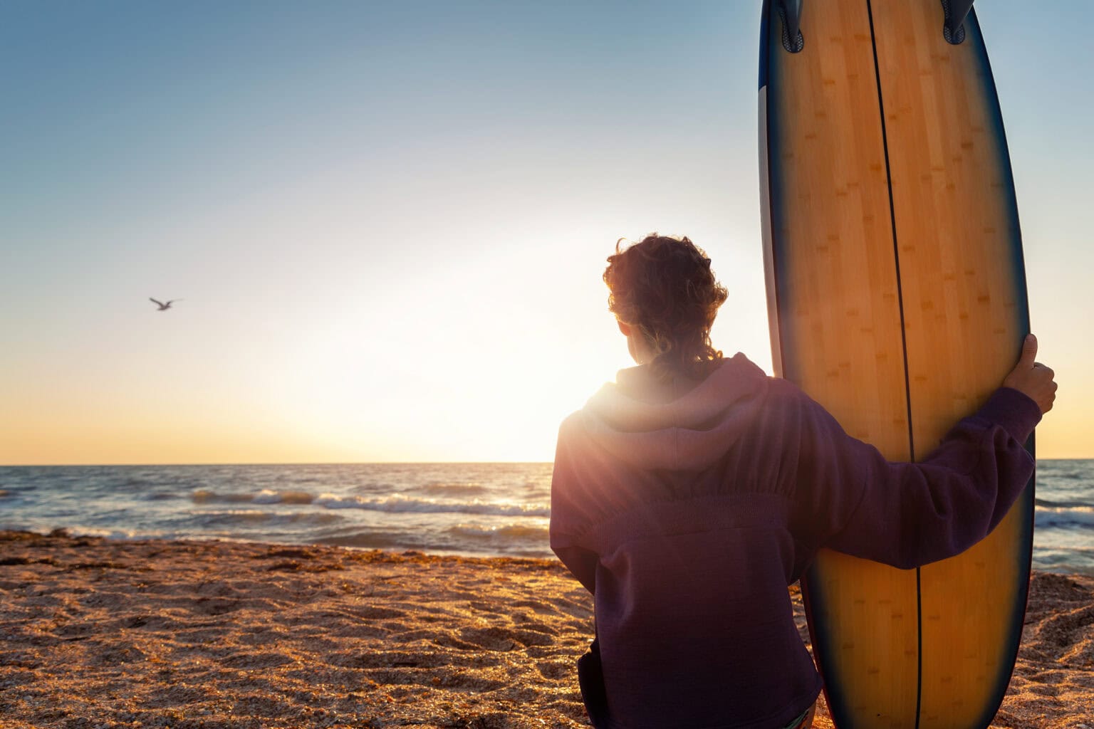 Back view young adult slim sporty female surfer girl with surfboard sitting on sand at ocean coast wave against warm sunrise or sunset sun